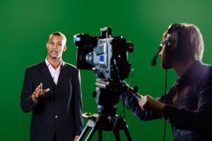 A television presenter in a TV studio with a camera and operator out of focus in the foreground and a green screen in the background. Some motion blur on the presenter's head and hands.